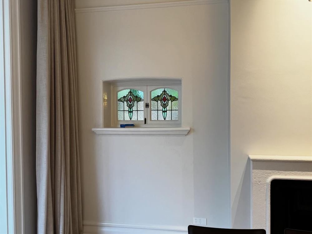 A leadlight window set into a white wall beside a curtain and mantel.