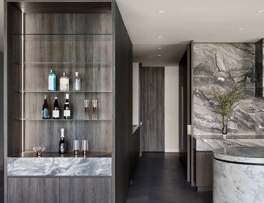 Bar with bottles on backlit shelving, marble surfaces and dark joinery.
