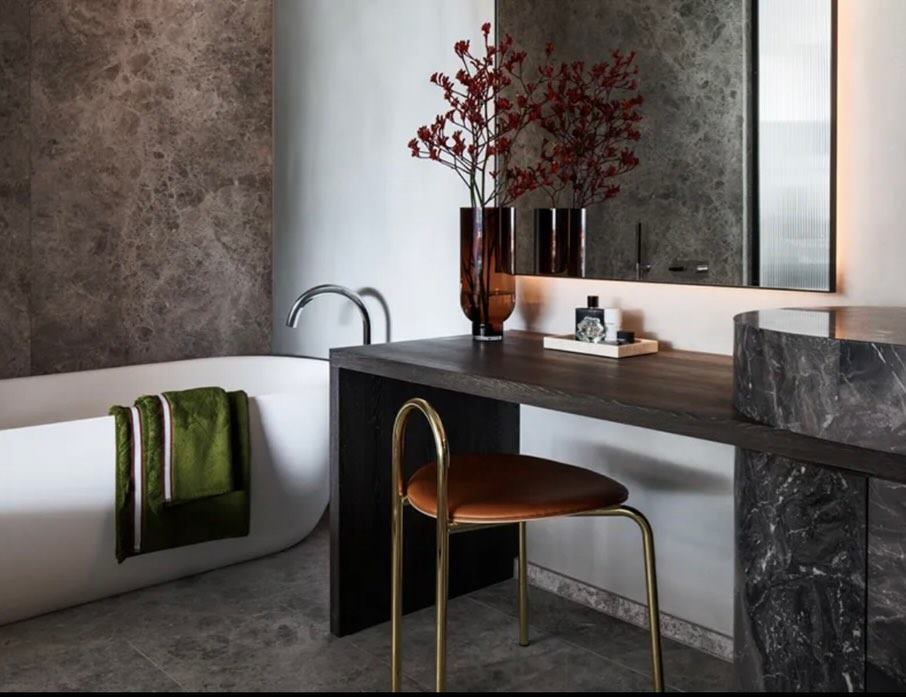 Bathroom with a freestanding bath, dark stone surfaces, a brass tap and a timber stool.