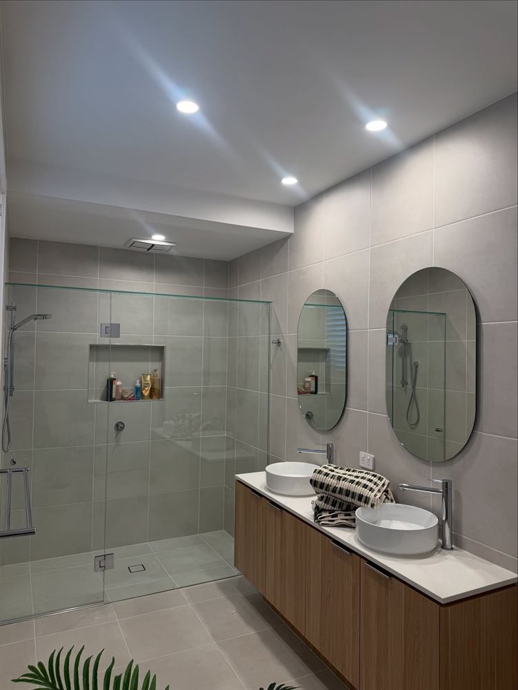Bathroom with a walk-in shower, timber vanity, two oval mirrors and pale grey tiling.