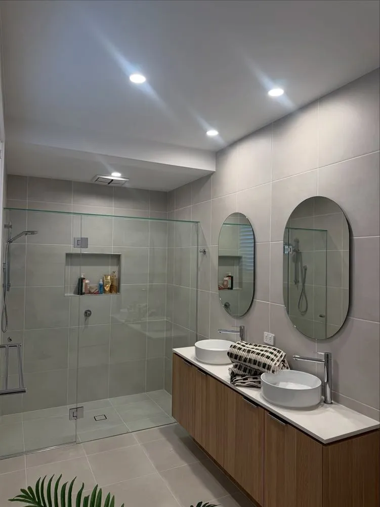 Bathroom with a walk-in shower, timber vanity, two oval mirrors and pale grey tiling.