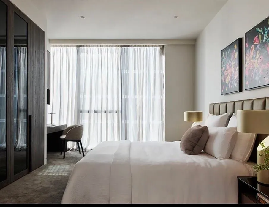 Bedroom with pale walls, full-height drapes, framed artwork and a writing desk.