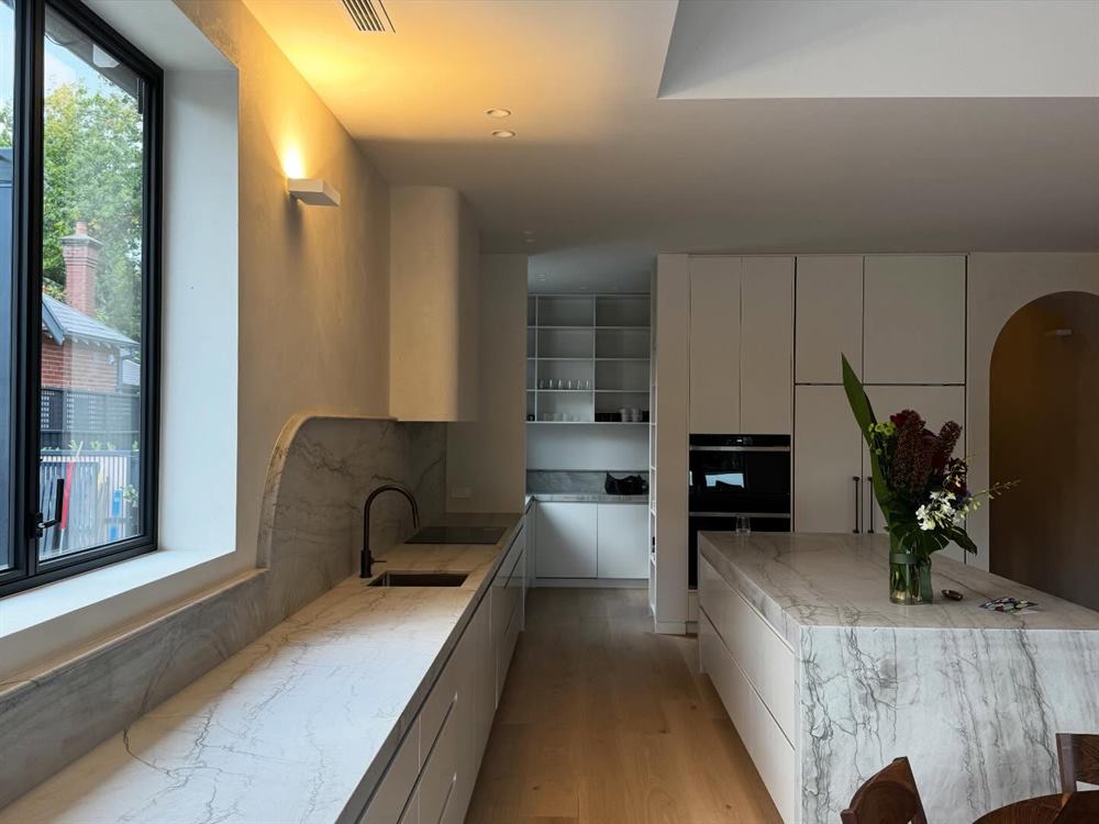 Kitchen with a marble island and splashback, an arched doorway and dark oven joinery.