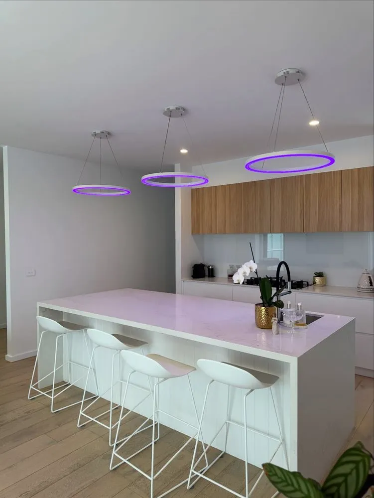 Kitchen with a white island bench, bar stools, timber joinery and violet ring pendant lights.