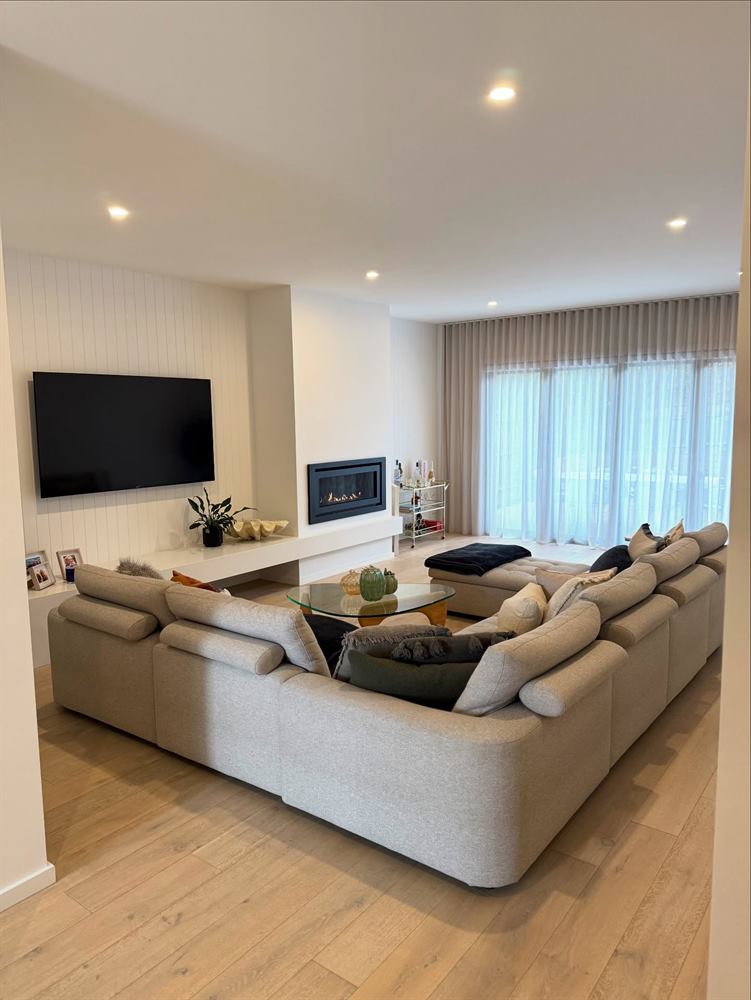 Living room with a large cream modular sofa, wall-mounted television, sheer curtains and timber floors.