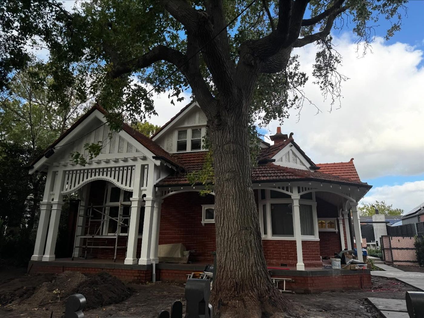 Edwardian red-brick home with white timber fretwork gables, mid-repaint beneath a large tree.