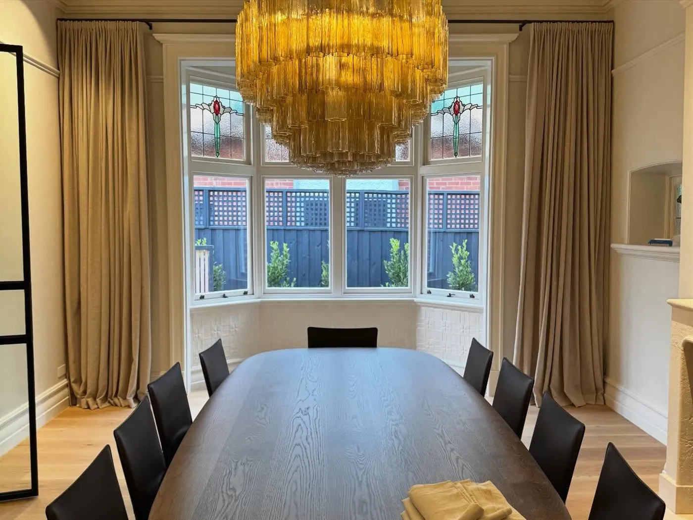 Dining room with an amber tiered chandelier, leadlight bay window and full-length curtains.