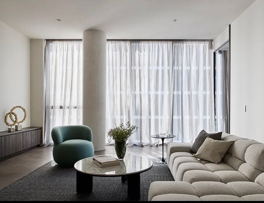 Living room with full-height sheer curtains, a pale rendered column, green armchair and low sofa.